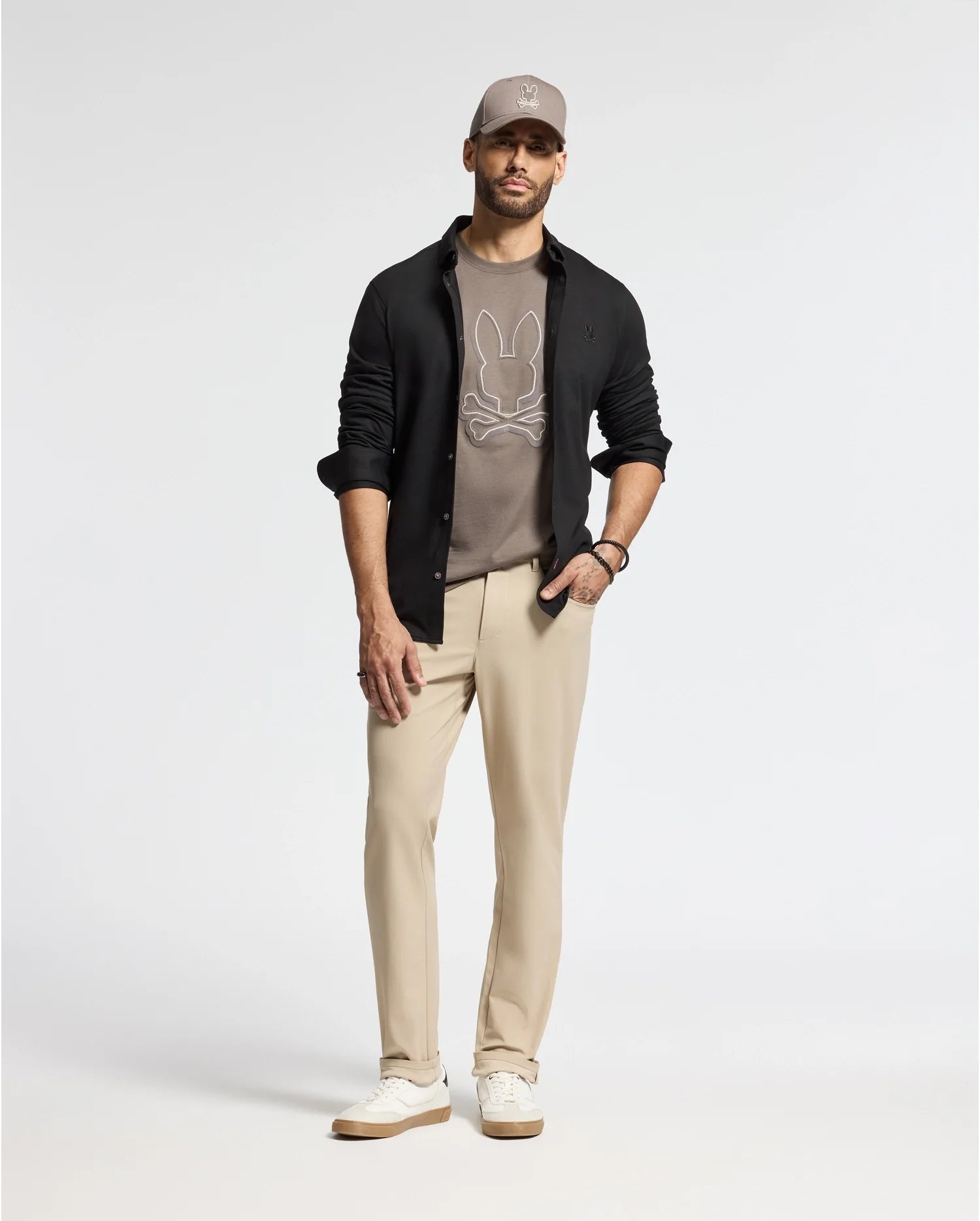 A man stands against a plain background, wearing a gray cap and an open black shirt. His outfit showcases the MENS ALEXANDER GRAPHIC TEE - B6U163E200 by Psycho Bunny, crafted from soft Pima cotton, paired with beige pants and white sneakers.