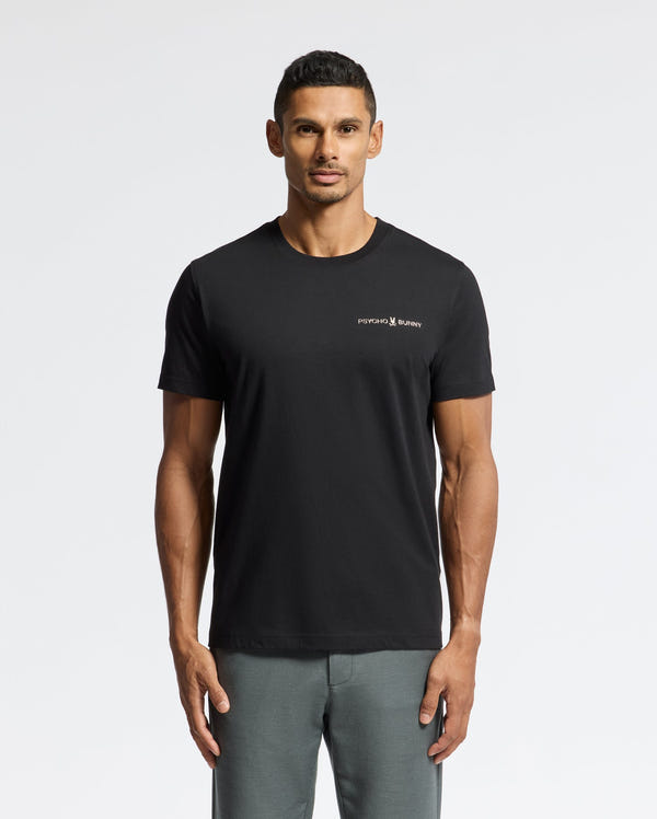A man is modeling the MENS LONGMONT GRAPHIC TEE - B6U194E200 by Psycho Bunny, featuring a stylish design with a small logo on the left chest. He stands against a plain white background, paired with gray pants, showcasing effortless style.