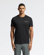 A man is modeling the MENS LONGMONT GRAPHIC TEE - B6U194E200 by Psycho Bunny, featuring a stylish design with a small logo on the left chest. He stands against a plain white background, paired with gray pants, showcasing effortless style.