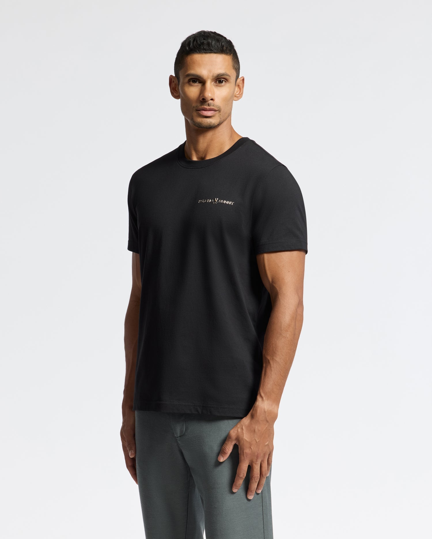 A man facing forward wears the stylish Psycho Bunny Men's Longmont Graphic Tee (B6U194E200), made from Pima cotton with white text, paired with gray pants against a plain white background.