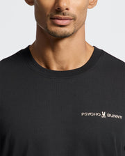 A person wearing the black "MEN'S LONGMONT GRAPHIC TEE - B6U194E200" by Psycho Bunny, a stylish Pima cotton graphic tee. The design features the brand's logo with a small bunny and skull motif on the chest. The background is plain and light-colored, perfectly showcasing this high-quality men's graphic tee.