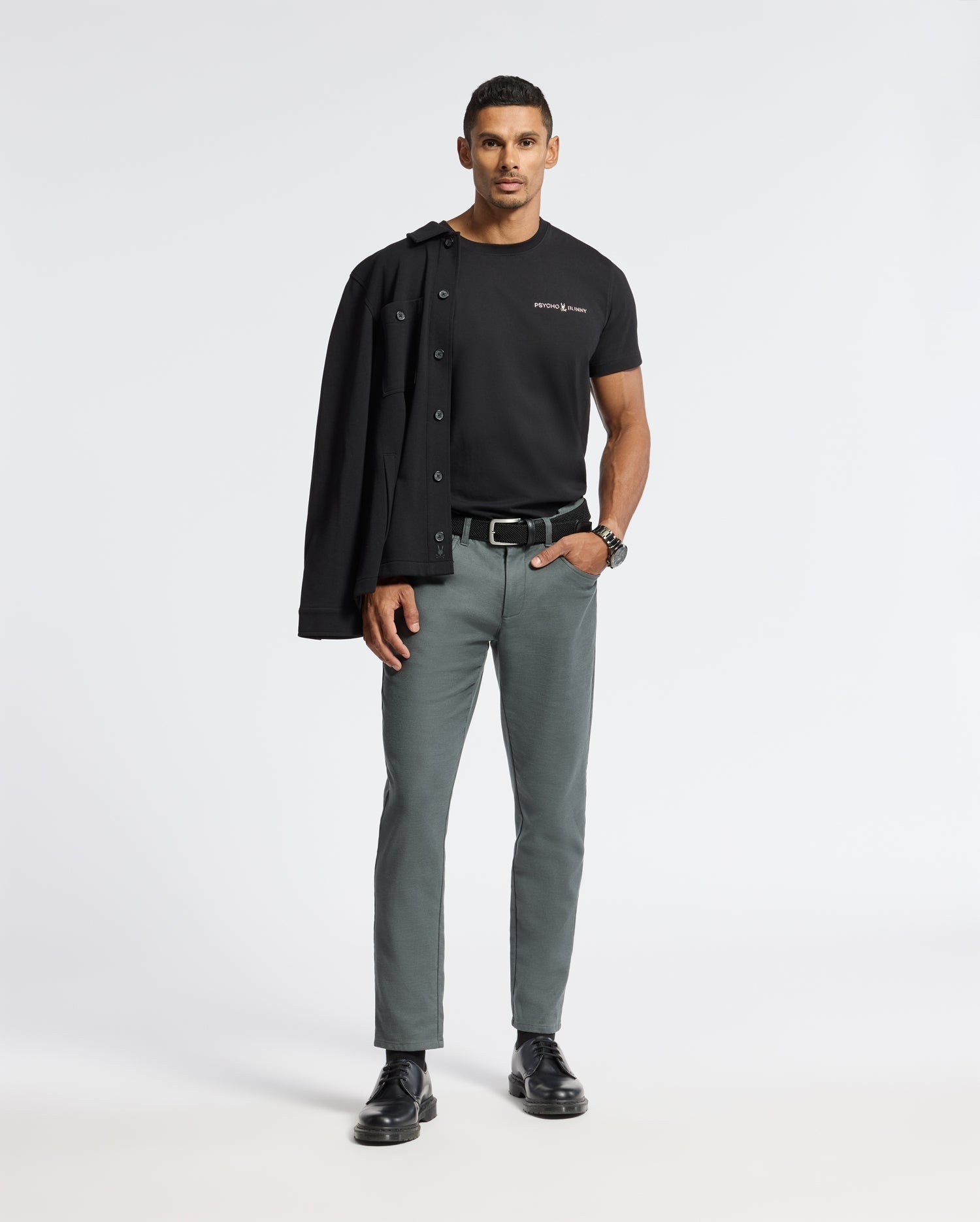 A man stands confidently against a plain background, wearing the stylish Psycho Bunny MENS LONGMONT GRAPHIC TEE - B6U194E200, paired with gray pants and black shoes. His black jacket is draped over his shoulder as he strikes a relaxed pose with one hand in his pocket.