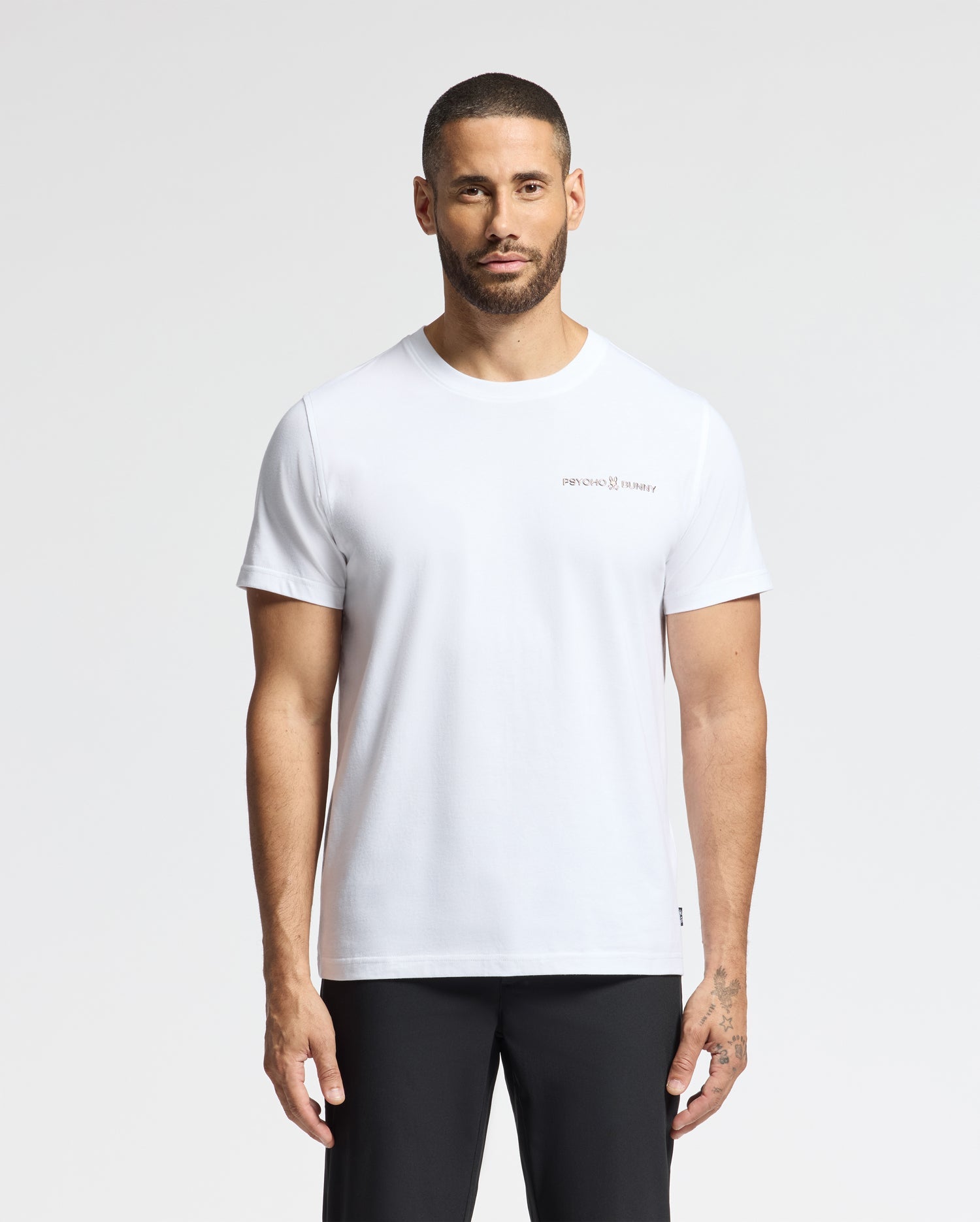 A man with short hair and a beard is wearing the Psycho Bunny MENS LONGMONT GRAPHIC TEE, crafted from soft Pima cotton and featuring a small logo on the chest. He pairs it with black pants as he stands against a light gray background.