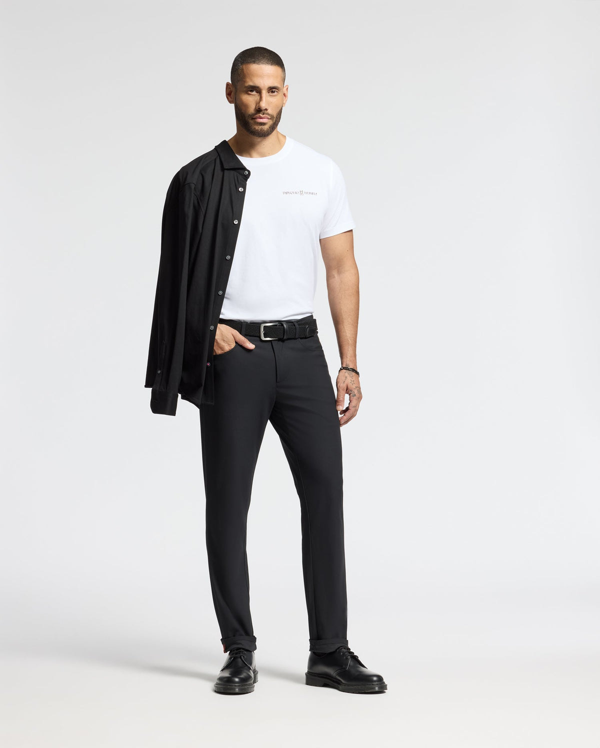 A man stands confidently, wearing a Psycho Bunny MENS LONGMONT GRAPHIC TEE - B6U194E200 in white, paired with black pants and shoes. Draped over his shoulder is a black shirt as he looks straight ahead against a plain white background.
