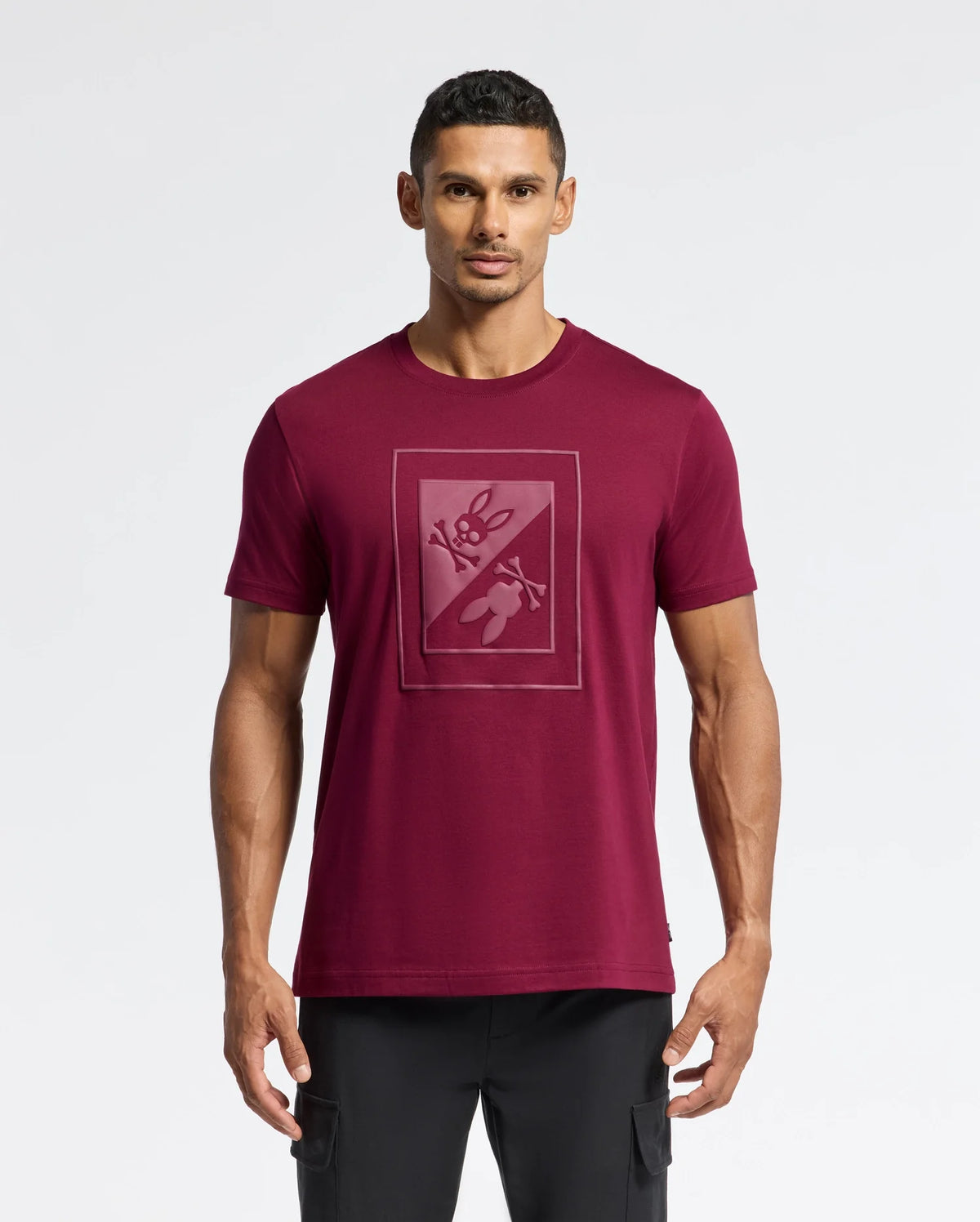 A man stands wearing a fashionable Psycho Bunny MENS DOTSERO GRAPHIC TEE - B6U197E200 in maroon, featuring a graphic design with paddles and a canoe, set against a plain white background.