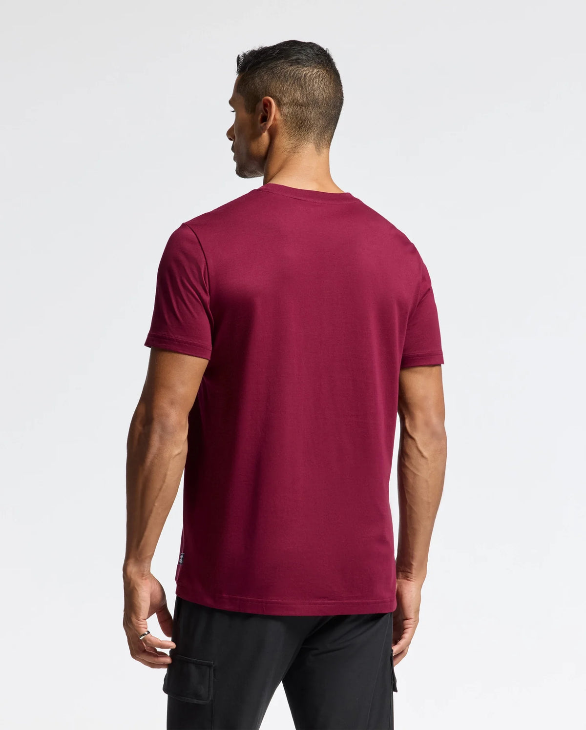 A person with short dark hair stands facing away from the camera, wearing a plain maroon MENS DOTSERO GRAPHIC TEE by Psycho Bunny and black pants against a white background.