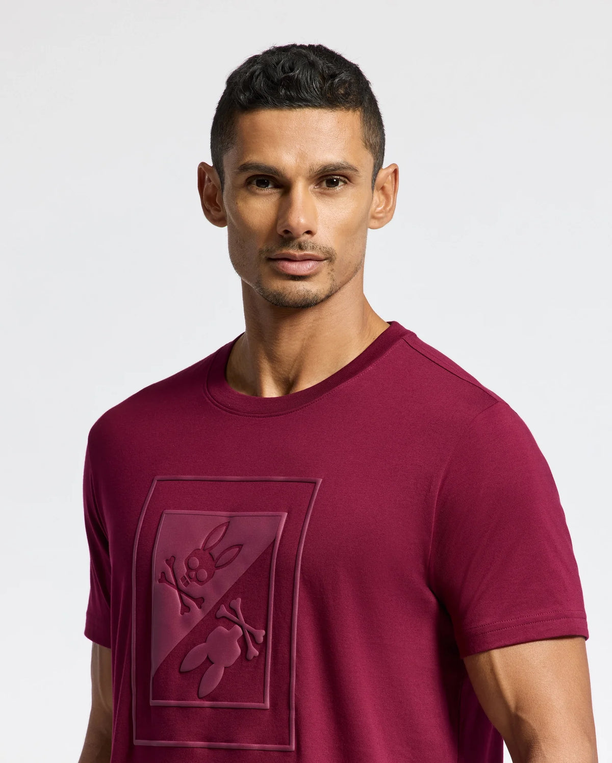 A man with short dark hair wears a stylish burgundy Psycho Bunny MENS DOTSERO GRAPHIC TEE (B6U197E200) showcasing a raised design of a cartoon character. The plain white background accentuates the allure of the graphic tee.