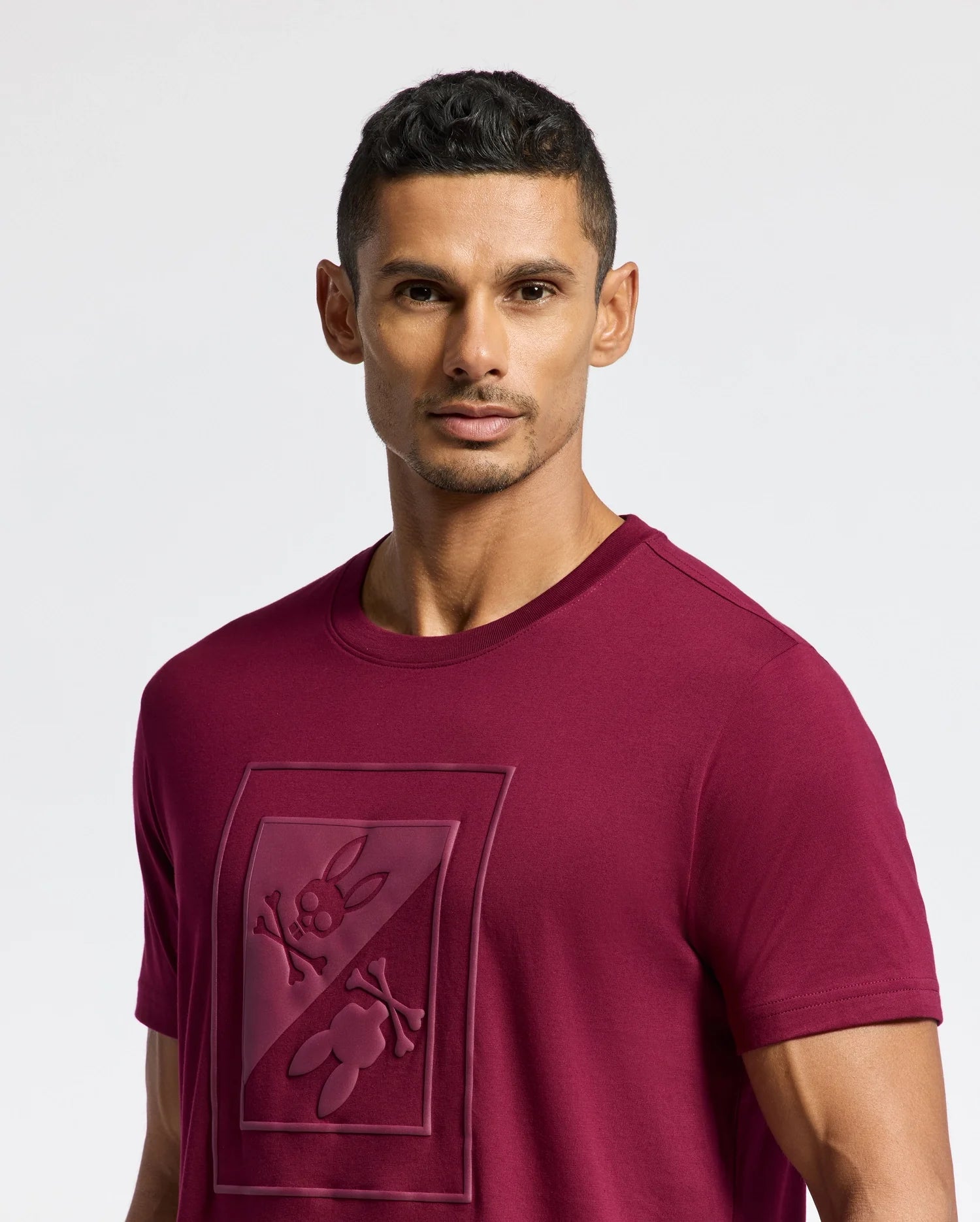 A man with short dark hair wears a stylish burgundy Psycho Bunny MENS DOTSERO GRAPHIC TEE (B6U197E200) showcasing a raised design of a cartoon character. The plain white background accentuates the allure of the graphic tee.