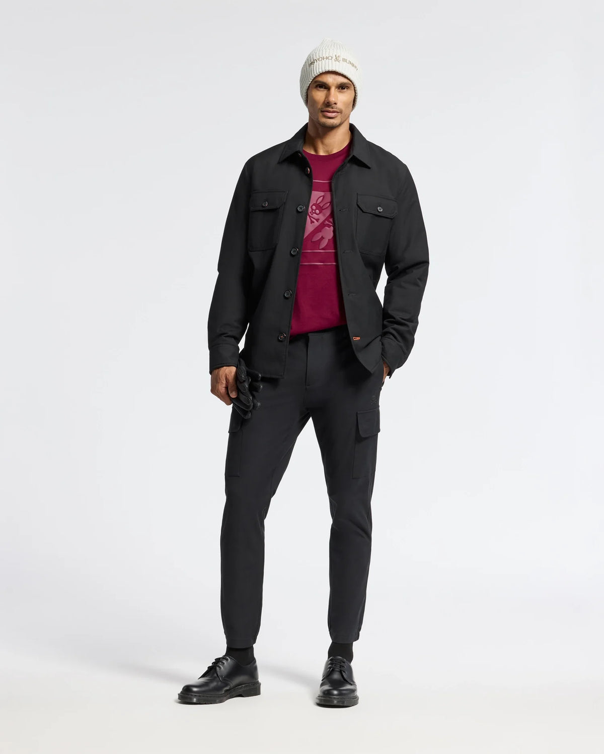 A man wearing a white beanie, black jacket, and a Psycho Bunny MENS DOTSERO GRAPHIC TEE in burgundy stands against a plain white background, complemented by black pants and shoes.