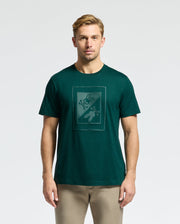 A man with short blonde hair is wearing a luxurious Psycho Bunny MENS DOTSERO GRAPHIC TEE - B6U197E200 in dark green, crafted from Peruvian Pima Cotton. He stands facing forward against a plain light background and pairs the tee with light brown pants.