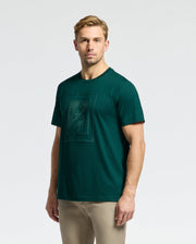 A person with short blond hair is wearing a Psycho Bunny MENS DOTSERO GRAPHIC TEE - B6U197E200 in dark green, featuring a square graphic on the front, paired with light beige pants. The background is plain white.