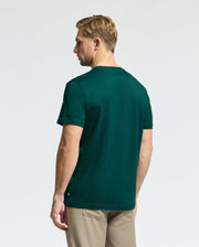 A man with short blonde hair is facing away from the camera, wearing a Psycho Bunny dark green MENS DOTSERO GRAPHIC TEE - B6U197E200 made from Peruvian Pima Cotton and light beige pants. The background is plain and light-colored.
