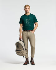 A man with short blond hair stands holding a beige backpack, wearing a Psycho Bunny men's Dotsero graphic tee in dark green, complemented by beige pants and brown boots. The background is plain white.
