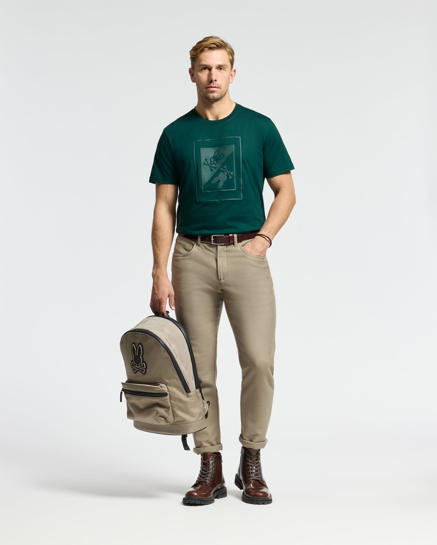 A man with short blond hair stands holding a beige backpack, wearing a Psycho Bunny men's Dotsero graphic tee in dark green, complemented by beige pants and brown boots. The background is plain white.