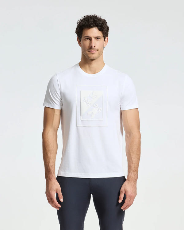 A man wearing a Psycho Bunny MENS DOTSER GRAPHIC TEE - B6U197E200, crafted from Pima cotton featuring a subtle embossed design on the front, pairs it with dark pants as he stands against a light gray background with his hands at his sides.