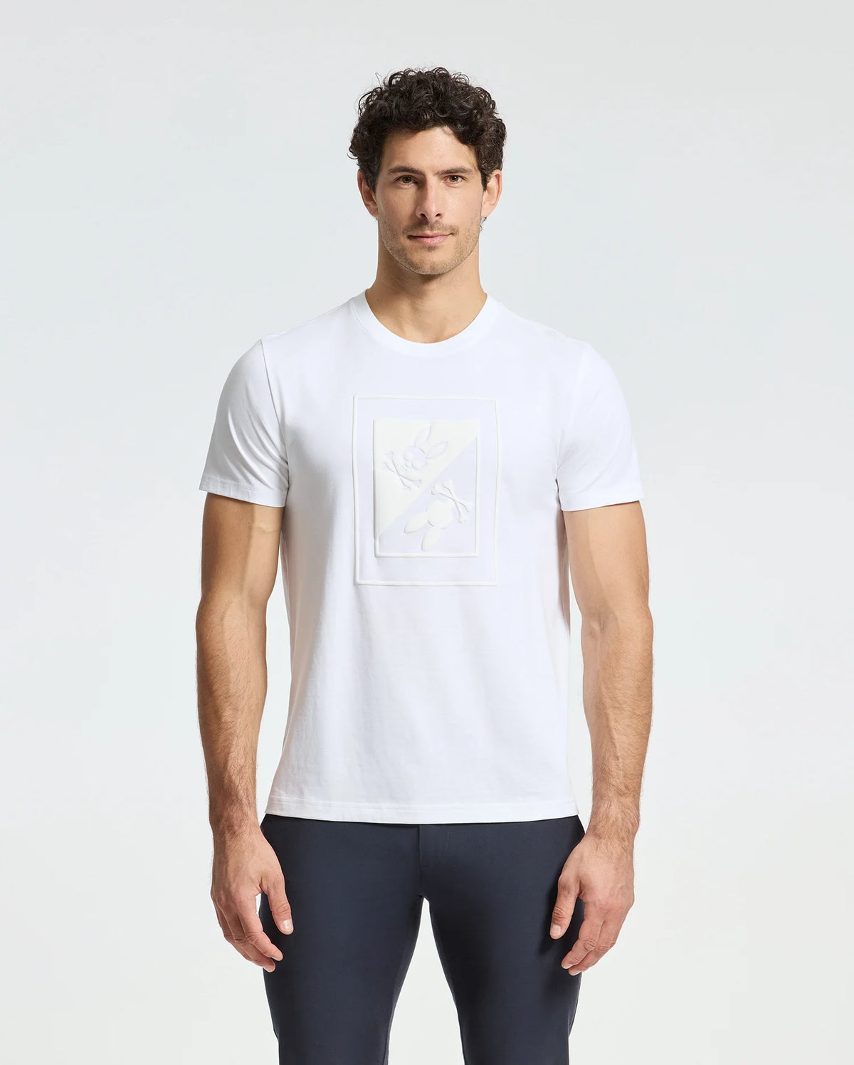 A man wearing a Psycho Bunny MENS DOTSER GRAPHIC TEE - B6U197E200, crafted from Pima cotton featuring a subtle embossed design on the front, pairs it with dark pants as he stands against a light gray background with his hands at his sides.