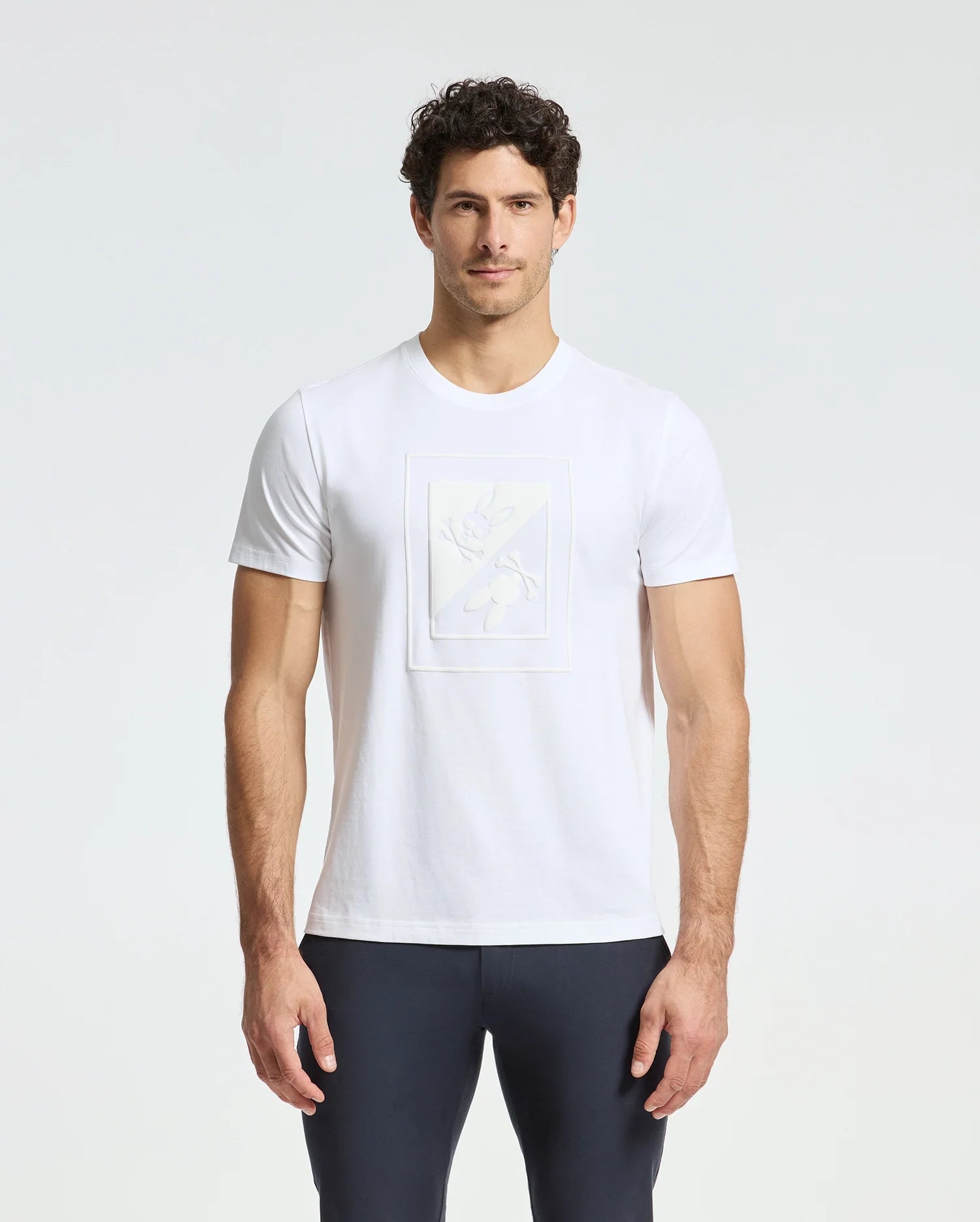 A man wearing a Psycho Bunny MENS DOTSER GRAPHIC TEE - B6U197E200, crafted from Pima cotton featuring a subtle embossed design on the front, pairs it with dark pants as he stands against a light gray background with his hands at his sides.