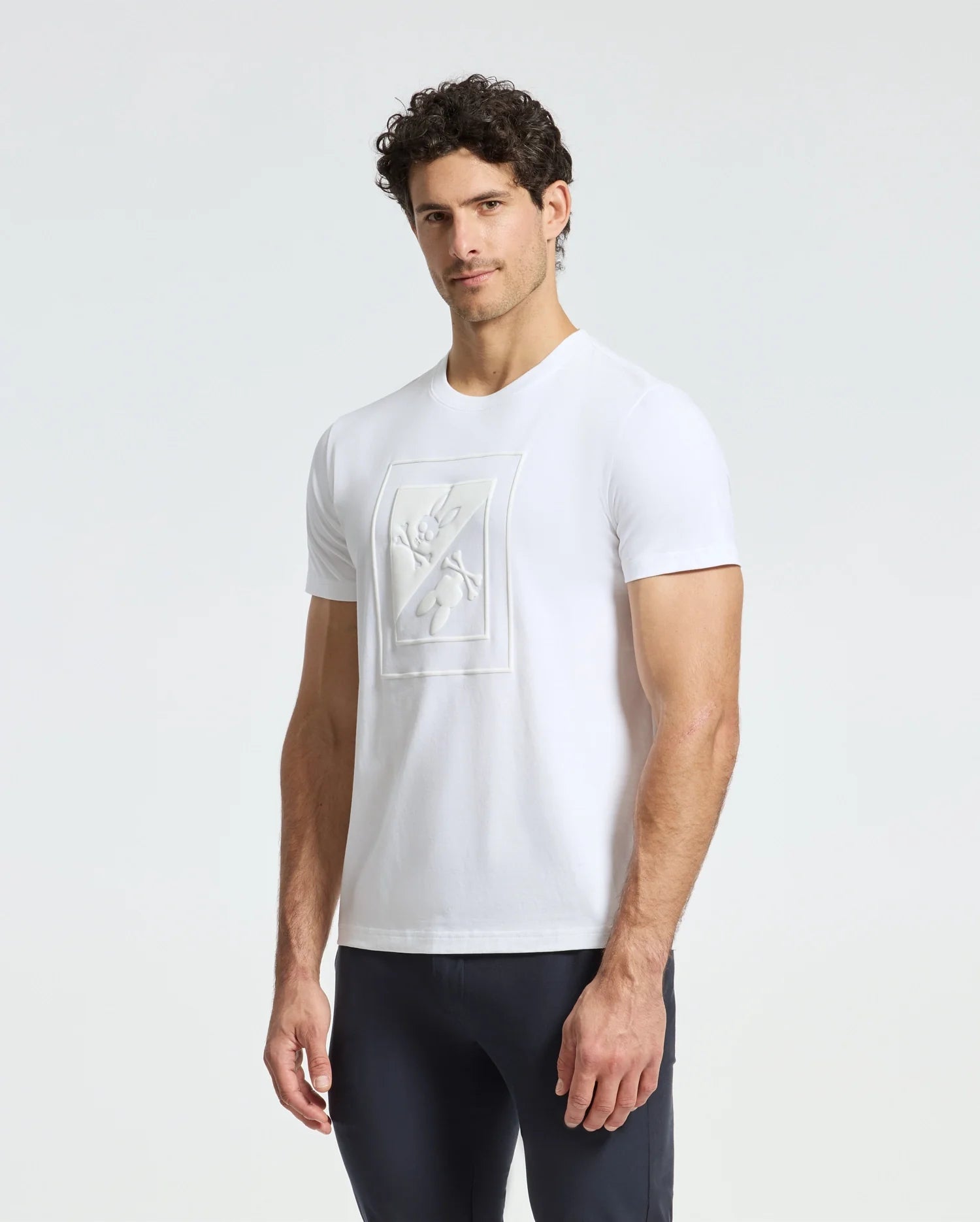 A man with curly hair is wearing a Psycho Bunny MENS DOTSER GRAPHIC TEE - B6U197E200, crafted from luxurious Peruvian Pima cotton, showcasing a textured design on the chest. He pairs it with dark pants and stands against a plain light-colored background.