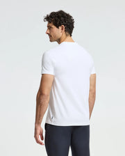 A man with curly hair is wearing a white Psycho Bunny MENS DOTSER GRAPHIC TEE - B6U197E200 and dark pants, standing and gazing to the side against a plain background.