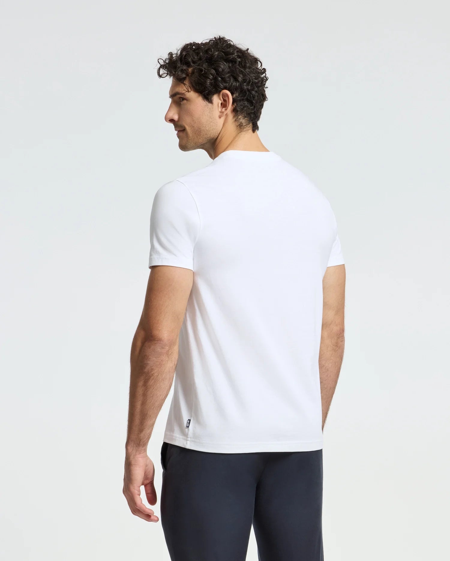 A man with curly hair is wearing a white Psycho Bunny MENS DOTSER GRAPHIC TEE - B6U197E200 and dark pants, standing and gazing to the side against a plain background.