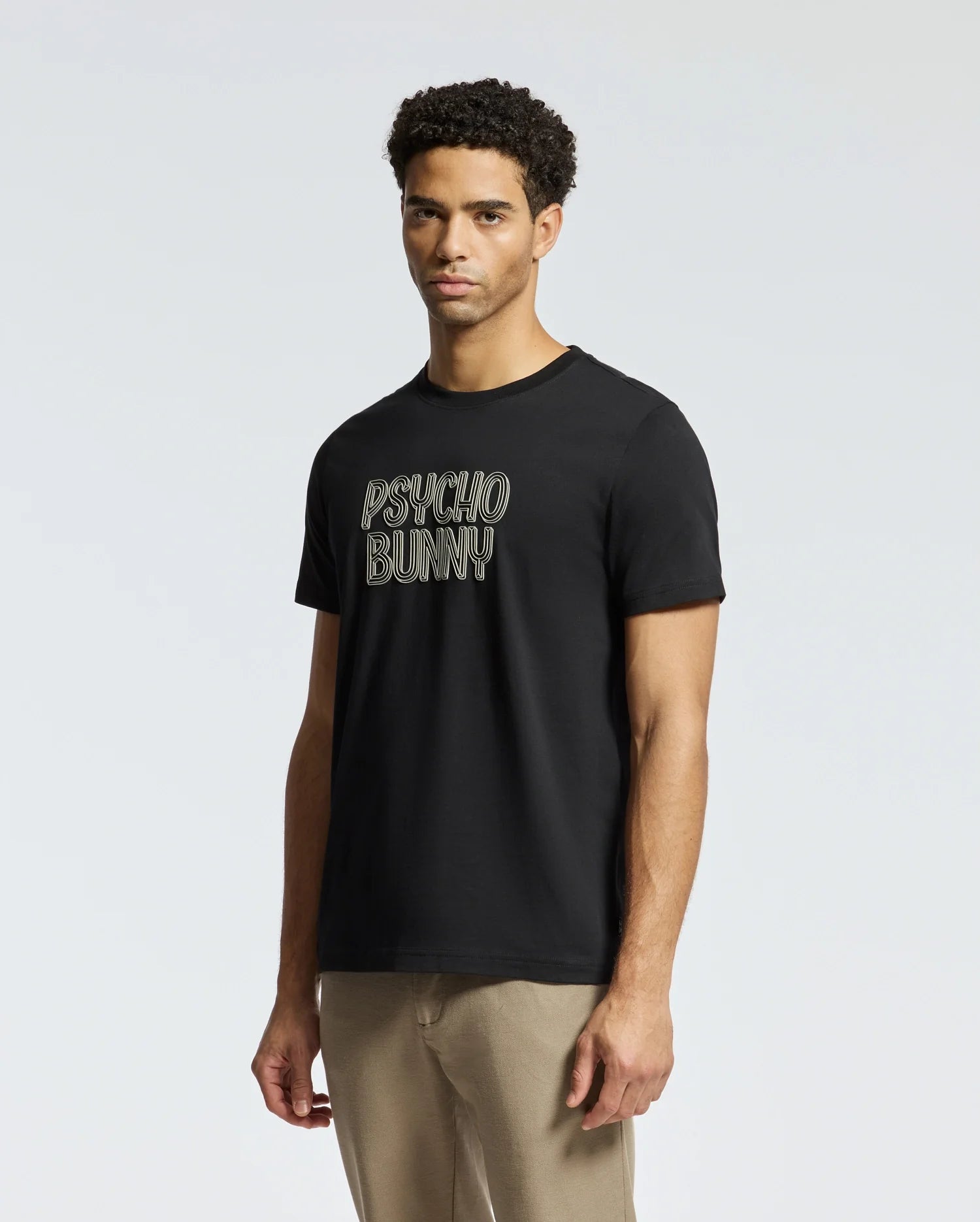 A person wears the MENS MONARC WORDMARK GRAPHIC TEE - B6U210E200 by Psycho Bunny in 100% Pima cotton, featuring the brand name "Psycho Bunny" against a plain background. Their curly hair complements the casual style of regular-fit beige pants.