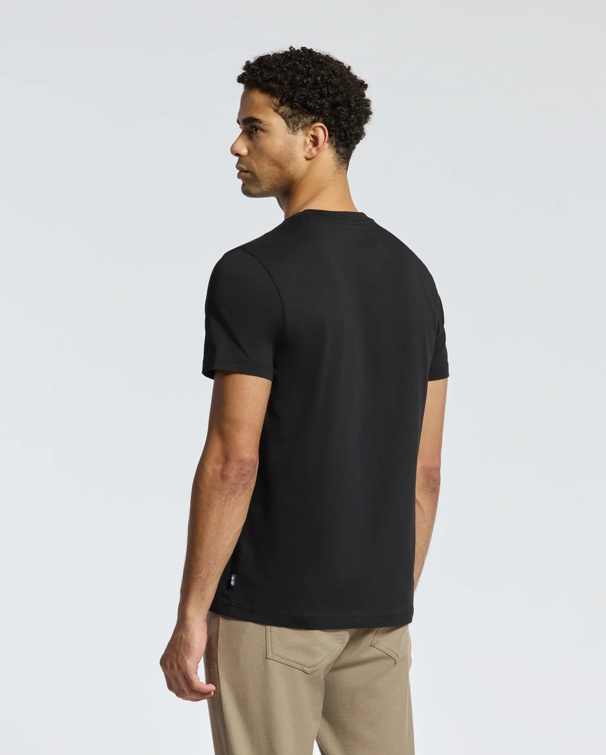 A man with short curly hair stands slightly turned, wearing a Psycho Bunny Mens Monarc Wordmark Graphic Tee crafted from 100% Pima cotton. His black T-shirt and beige pants have a regular fit. The background is plain and light-colored.