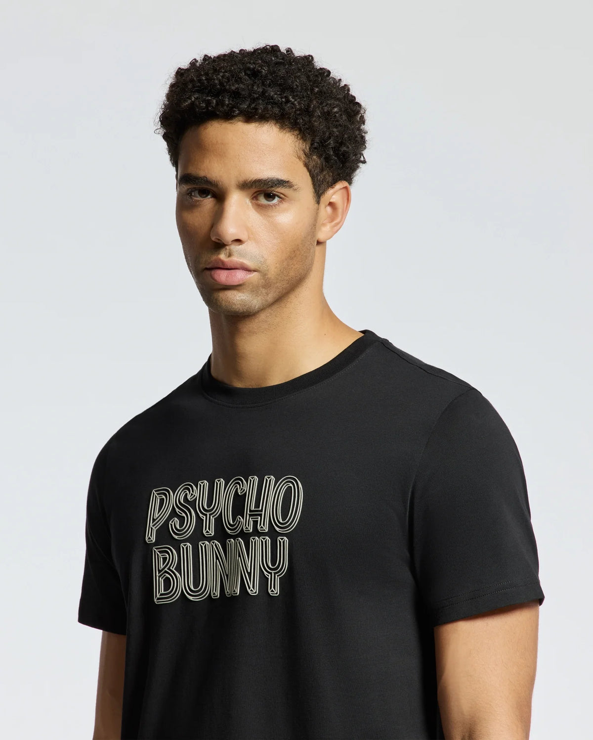 A person with short curly hair wearing the MENS MONARC WORDMARK GRAPHIC TEE - B6U210E200 in a regular fit black design by Psycho Bunny stands against a plain, light gray background. The Tee, made from 100% Pima cotton, effortlessly complements their neutral expression.