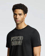 A person with short curly hair wearing the MENS MONARC WORDMARK GRAPHIC TEE - B6U210E200 in a regular fit black design by Psycho Bunny stands against a plain, light gray background. The Tee, made from 100% Pima cotton, effortlessly complements their neutral expression.