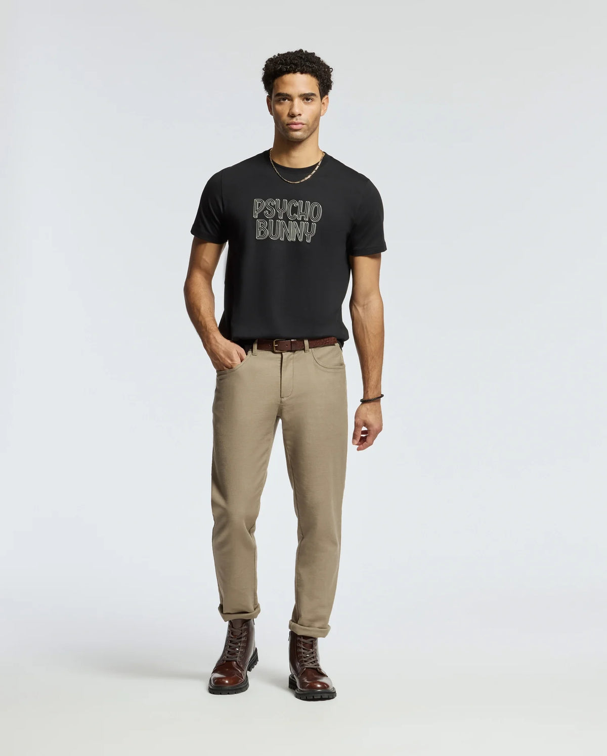 A man stands against a plain background, wearing the MEN'S MONARC WORDMARK GRAPHIC TEE - B6U210E200 from Psycho Bunny. The regular fit T-shirt, crafted from 100% Pima cotton, is in black and is paired with beige pants and dark brown boots. His hands rest calmly by his sides.