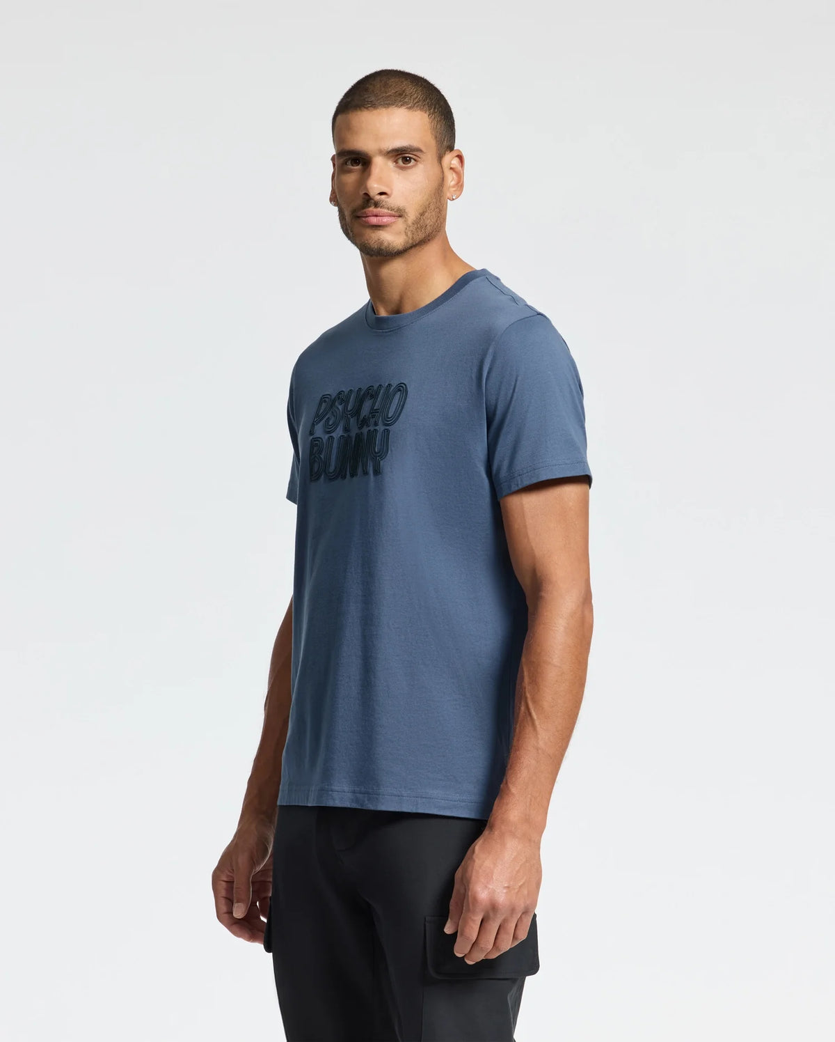 A man in a blue "MENS MONARC WORDMARK GRAPHIC TEE - B6U210E200" by Psycho Bunny, crafted from soft Pima cotton, stands comfortably against a plain white background, with his black pants providing a classic touch.