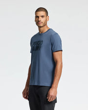 A man in a blue "MENS MONARC WORDMARK GRAPHIC TEE - B6U210E200" by Psycho Bunny, crafted from soft Pima cotton, stands comfortably against a plain white background, with his black pants providing a classic touch.