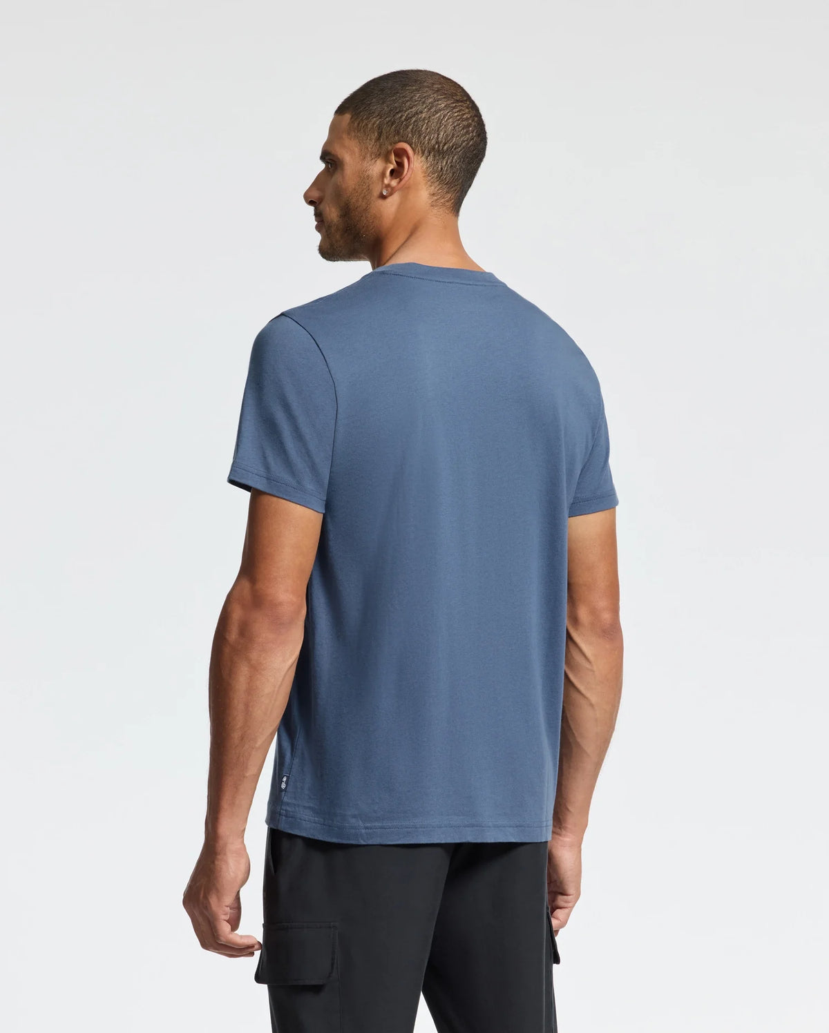 A man with short hair faces away from the camera, wearing a Psycho Bunny MENS MONARC WORDMARK GRAPHIC TEE crafted from soft Pima cotton for optimal comfort, complemented by black pants. The background is a plain white.