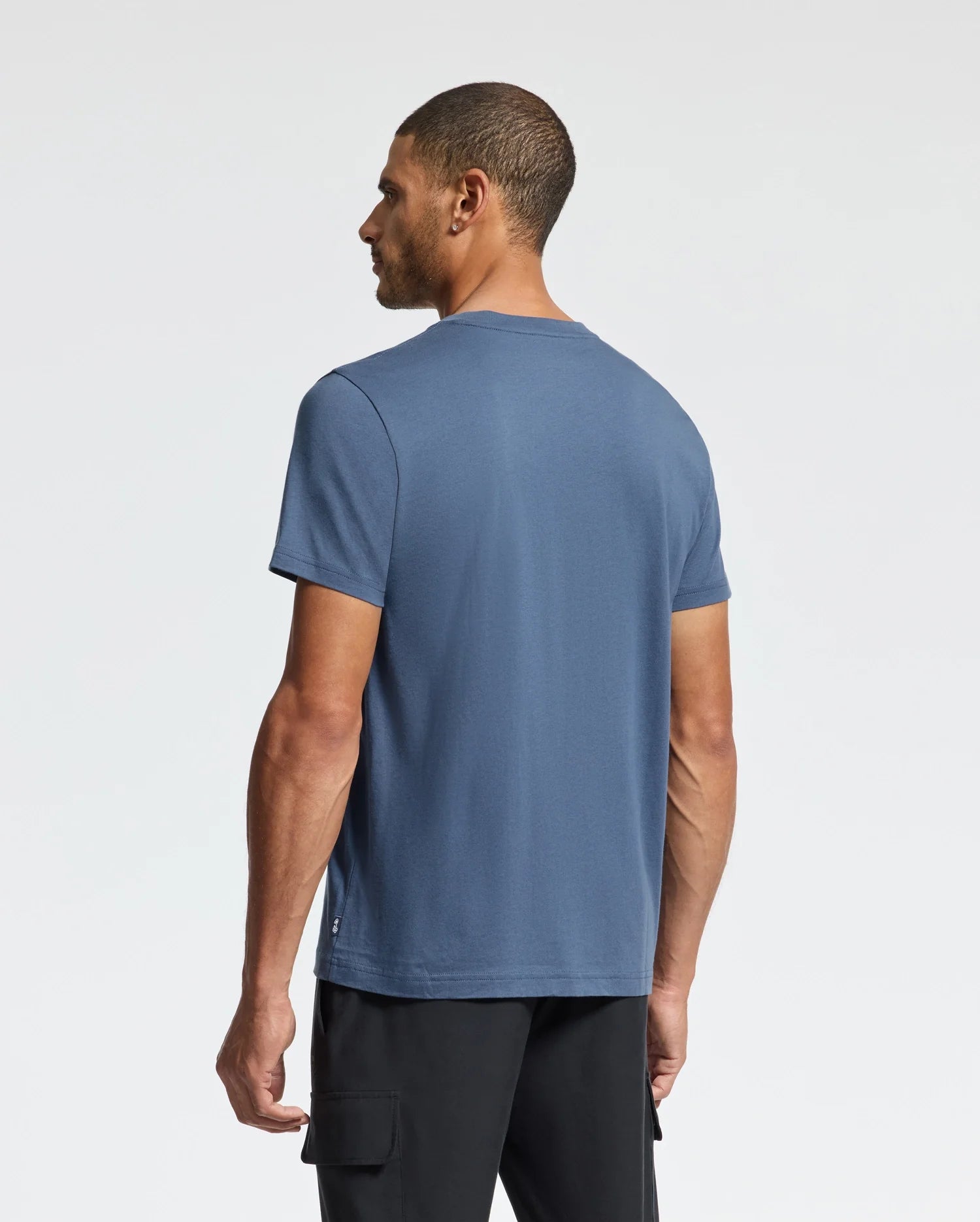 A man with short hair faces away from the camera, wearing a Psycho Bunny MENS MONARC WORDMARK GRAPHIC TEE crafted from soft Pima cotton for optimal comfort, complemented by black pants. The background is a plain white.