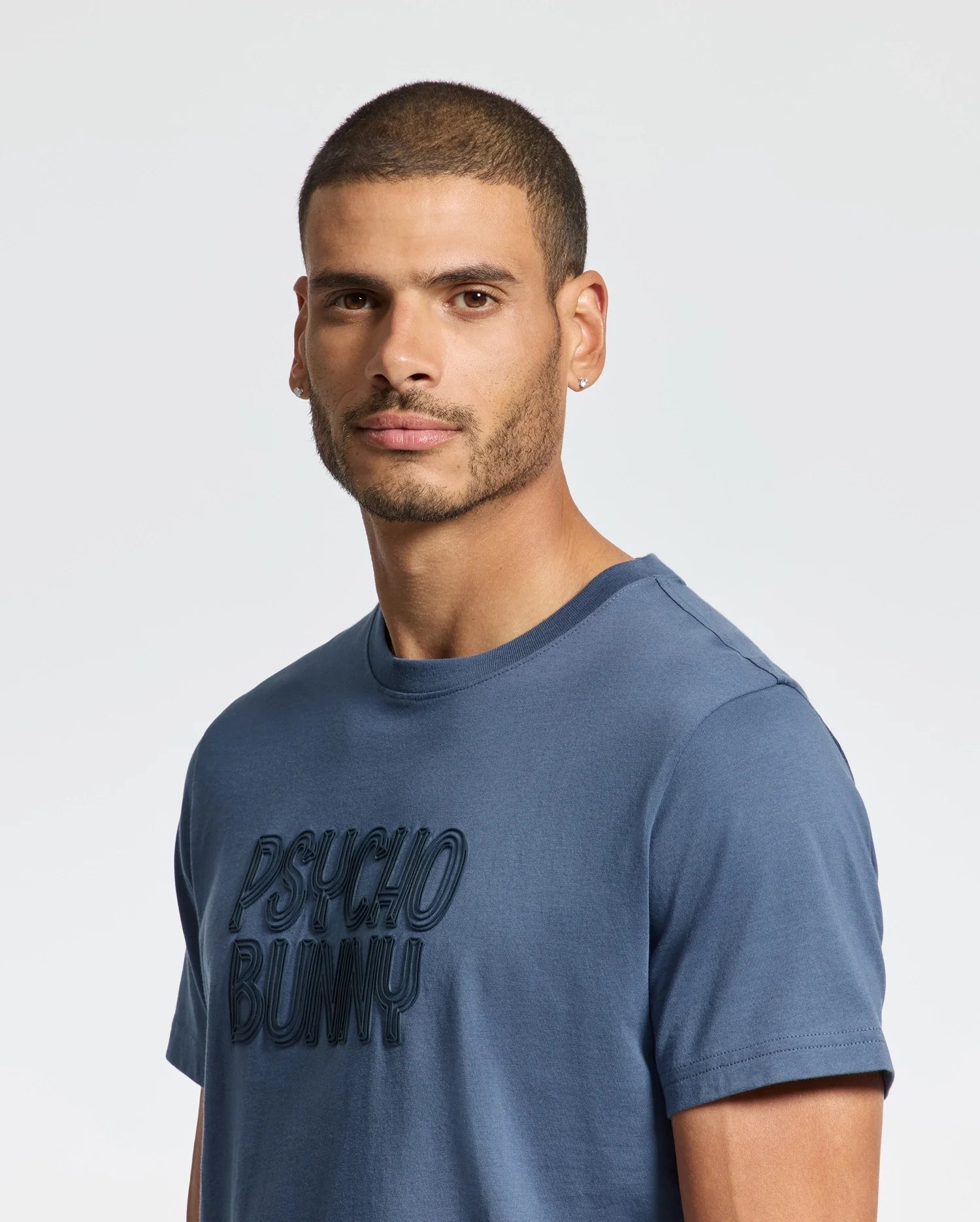 A man with short hair and a beard is wearing a blue MENS MONARC WORDMARK GRAPHIC TEE - B6U210E200 by Psycho Bunny, crafted from soft Pima cotton. He has a neutral expression, and the background is an off-white color.