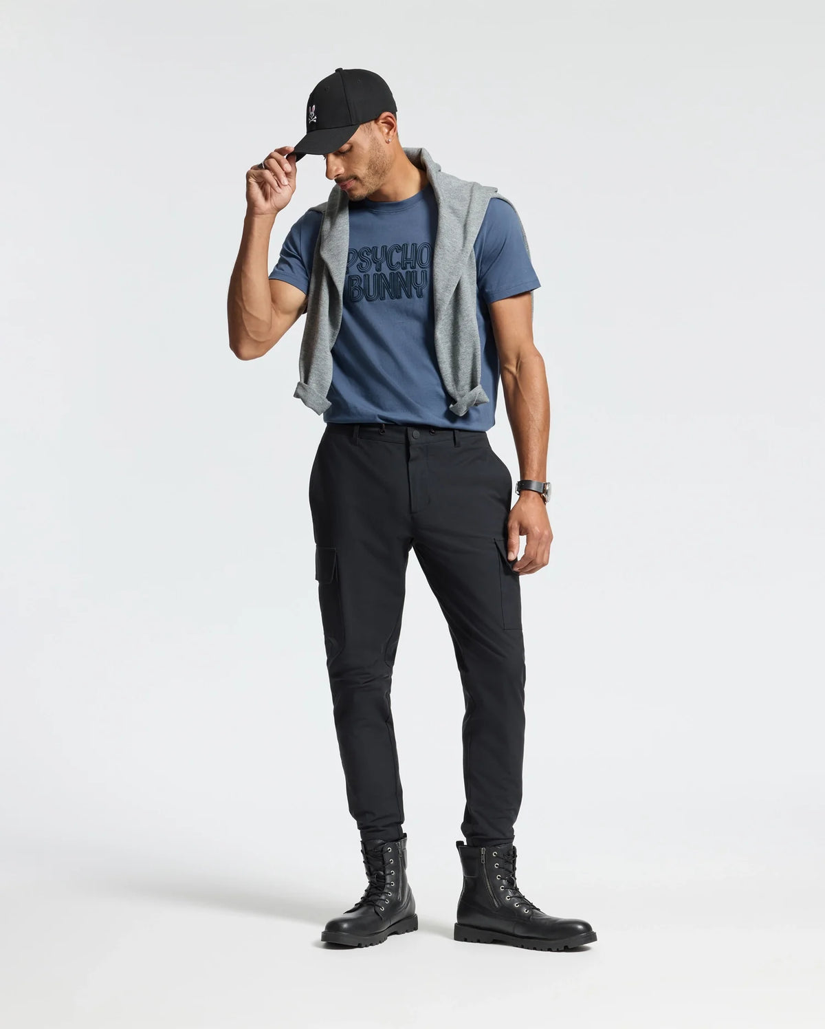 A person stands against a plain background wearing a cap, the blue Psycho Bunny Mens Monarc Wordmark Graphic Tee (B6U210E200), black pants, and boots. A gray sweater is draped over their shoulders for comfort, and they are looking down, holding the brim of the cap with one hand.