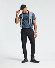 A person stands against a plain background wearing a cap, the blue Psycho Bunny Mens Monarc Wordmark Graphic Tee (B6U210E200), black pants, and boots. A gray sweater is draped over their shoulders for comfort, and they are looking down, holding the brim of the cap with one hand.