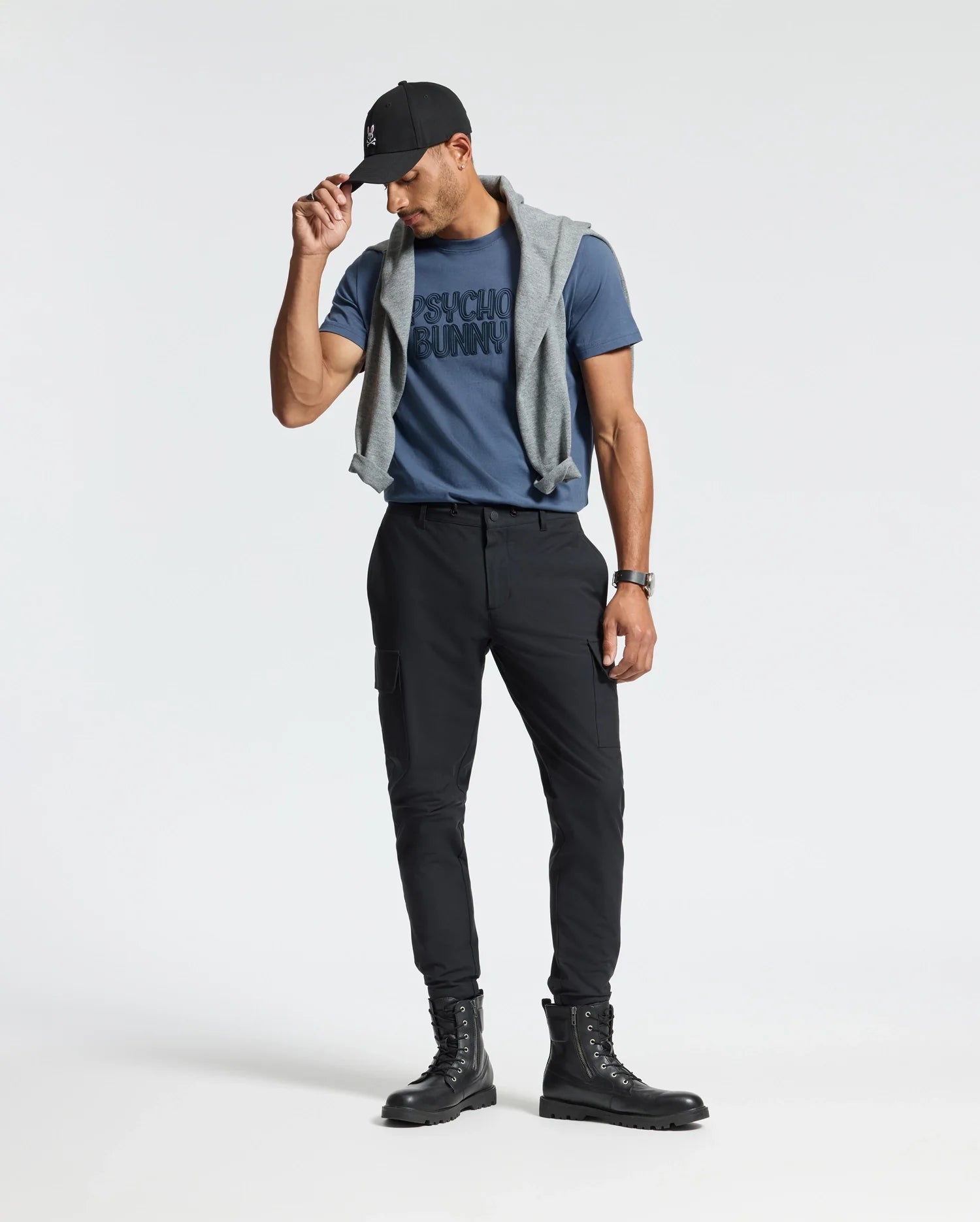 A person stands against a plain background wearing a cap, the blue Psycho Bunny Mens Monarc Wordmark Graphic Tee (B6U210E200), black pants, and boots. A gray sweater is draped over their shoulders for comfort, and they are looking down, holding the brim of the cap with one hand.