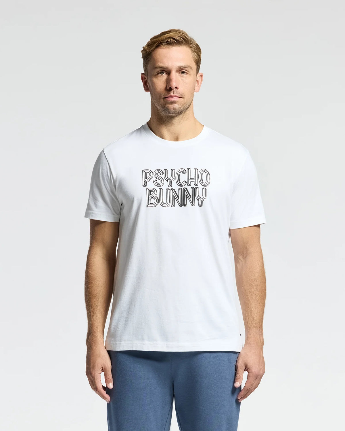 A man stands wearing the MENS MONARC WORDMARK GRAPHIC TEE - B6U210E200 from Psycho Bunny, crafted from premium Pima cotton with "Psycho Bunny" printed on it, paired with blue pants. Made in Peru, the tee features a plain, light background for stylish simplicity.