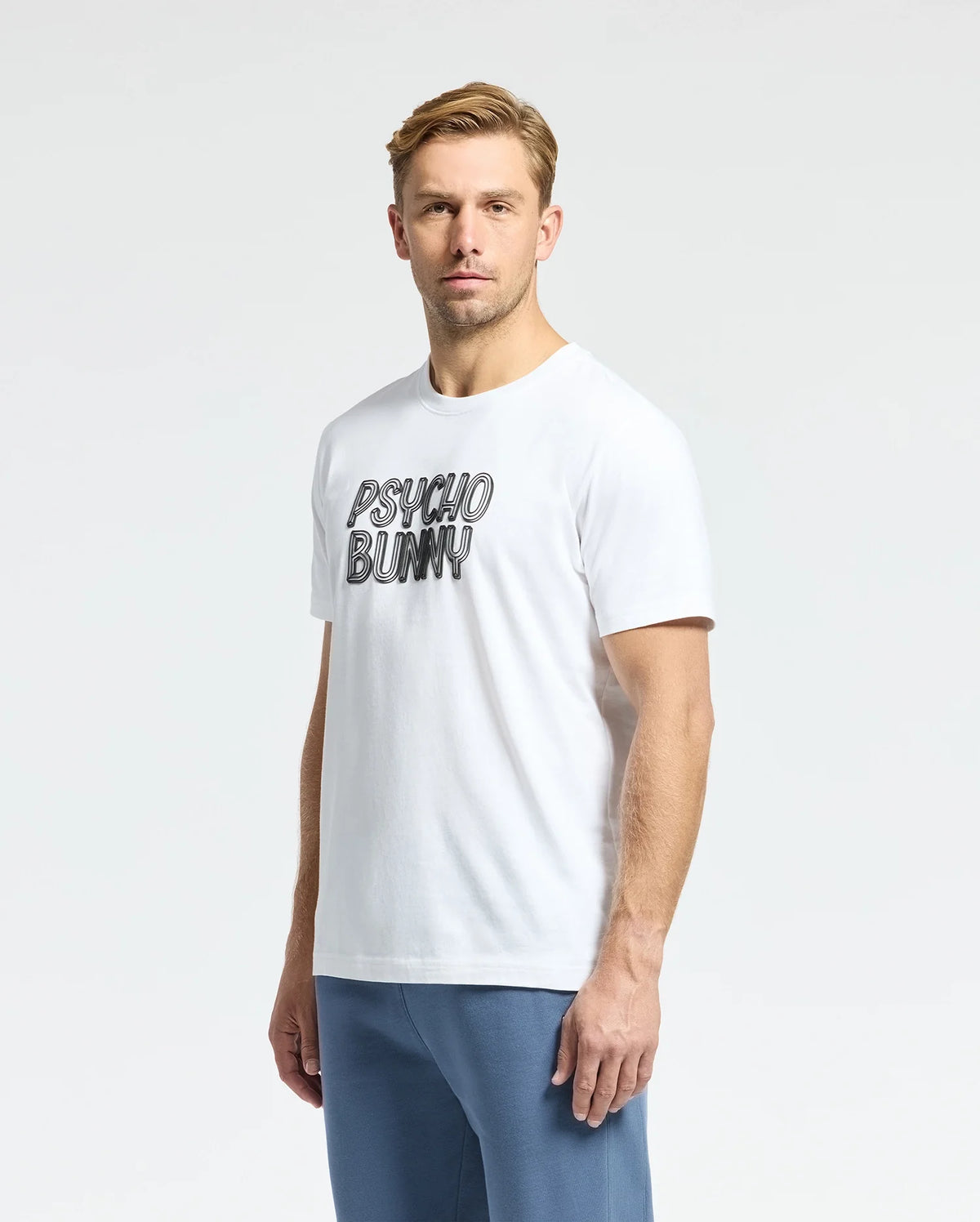 A man with short hair stands against a plain white background, wearing the Psycho Bunny Men's Monarc Wordmark Graphic Tee (B6U210E200), crafted from luxurious Pima cotton in Peru. The crisp, white T-shirt features bold "Psycho Bunny" lettering and is paired stylishly with blue pants.