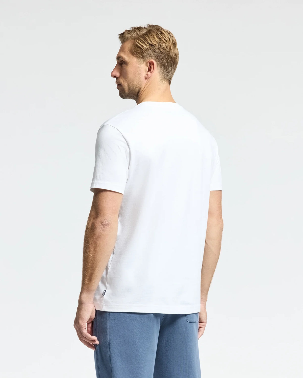A man with short blond hair is facing away from the camera, wearing Psycho Bunny’s MENS MONARC WORDMARK GRAPHIC TEE in white Pima cotton and blue pants, set against a simple, light-colored background.