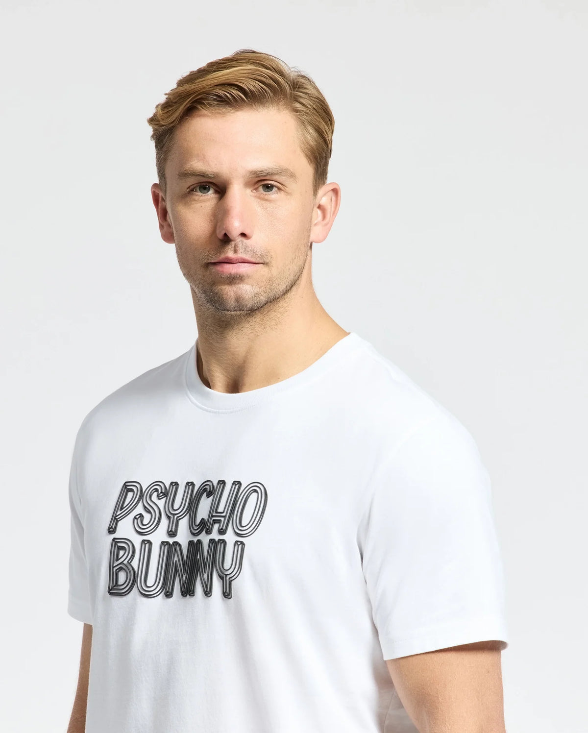 A person with short blond hair wears a white Psycho Bunny Monarc Wordmark Graphic Tee, made from premium Pima cotton, featuring the "PSYCHO BUNNY" print. The simple background is plain white.