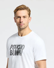 A person with short blond hair wears a white Psycho Bunny Monarc Wordmark Graphic Tee, made from premium Pima cotton, featuring the "PSYCHO BUNNY" print. The simple background is plain white.