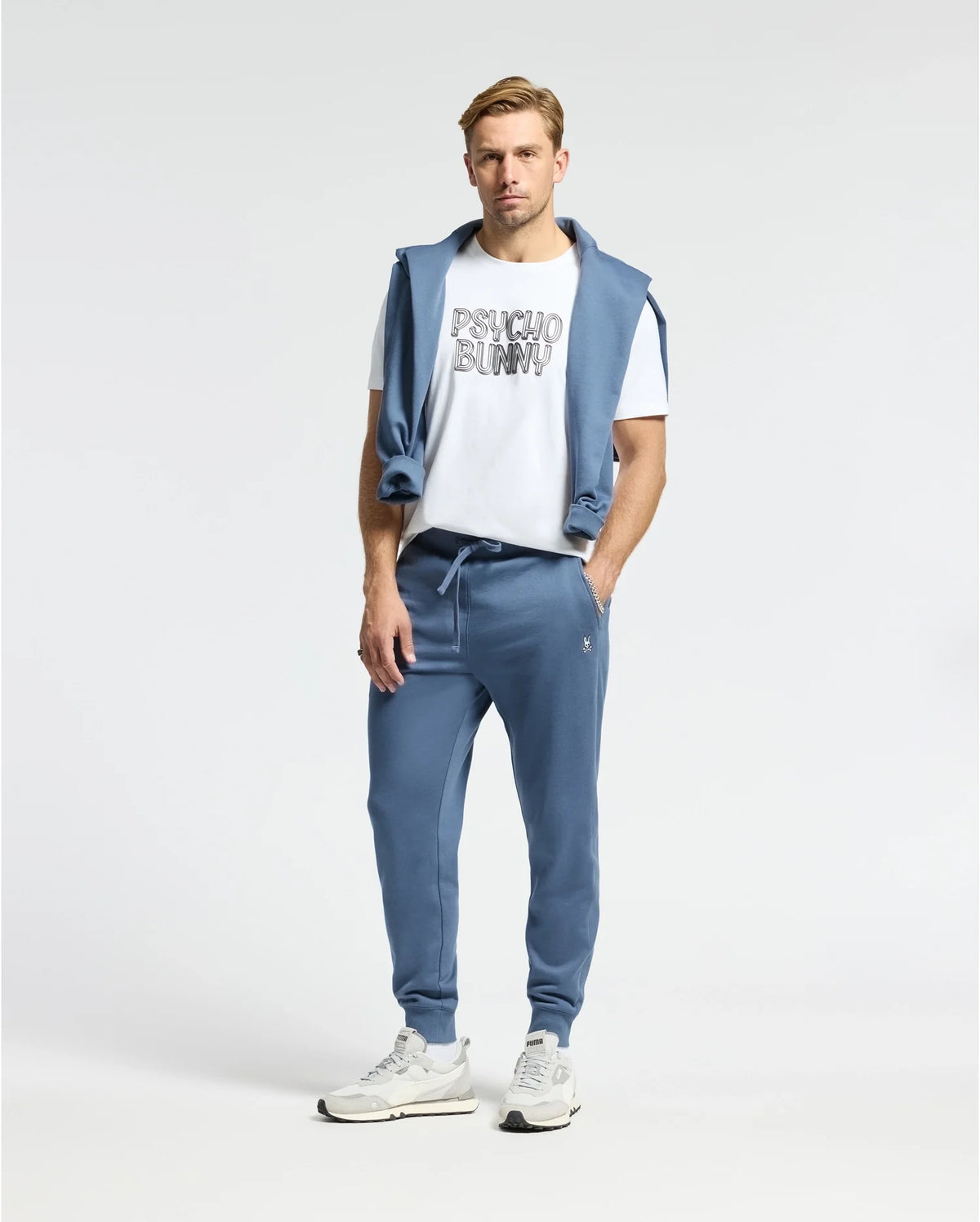 A man stands relaxed in a white Psycho Bunny Men's Monarc Wordmark Graphic Tee, with a blue sweater over his shoulders and matching jogger pants. He completes the ensemble with white sneakers against a plain white background.
