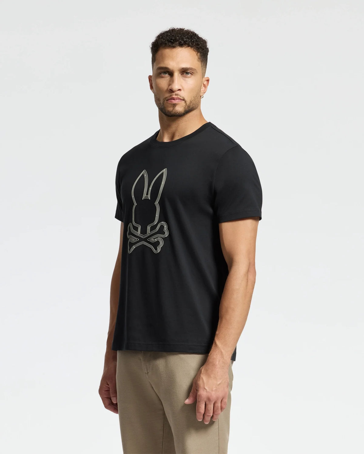 A man wearing the Psycho Bunny MENS MONARC FRONT BUNNY GRAPHIC TEE - B6U211E200, featuring a rib knit crewneck and crafted from 100% Pima cotton for ultimate comfort, stands against a plain white background. He pairs it perfectly with beige pants.