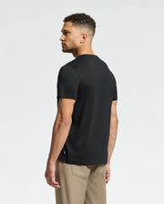 A man with short curly hair and a trimmed beard stands sideways, showcasing a black Psycho Bunny Men's Monarc Front Bunny Graphic Tee (B6U211E200), crafted from 100% Pima cotton. He pairs it with beige pants, while the rib knit crewneck provides an excellent contrast to the plain white background.