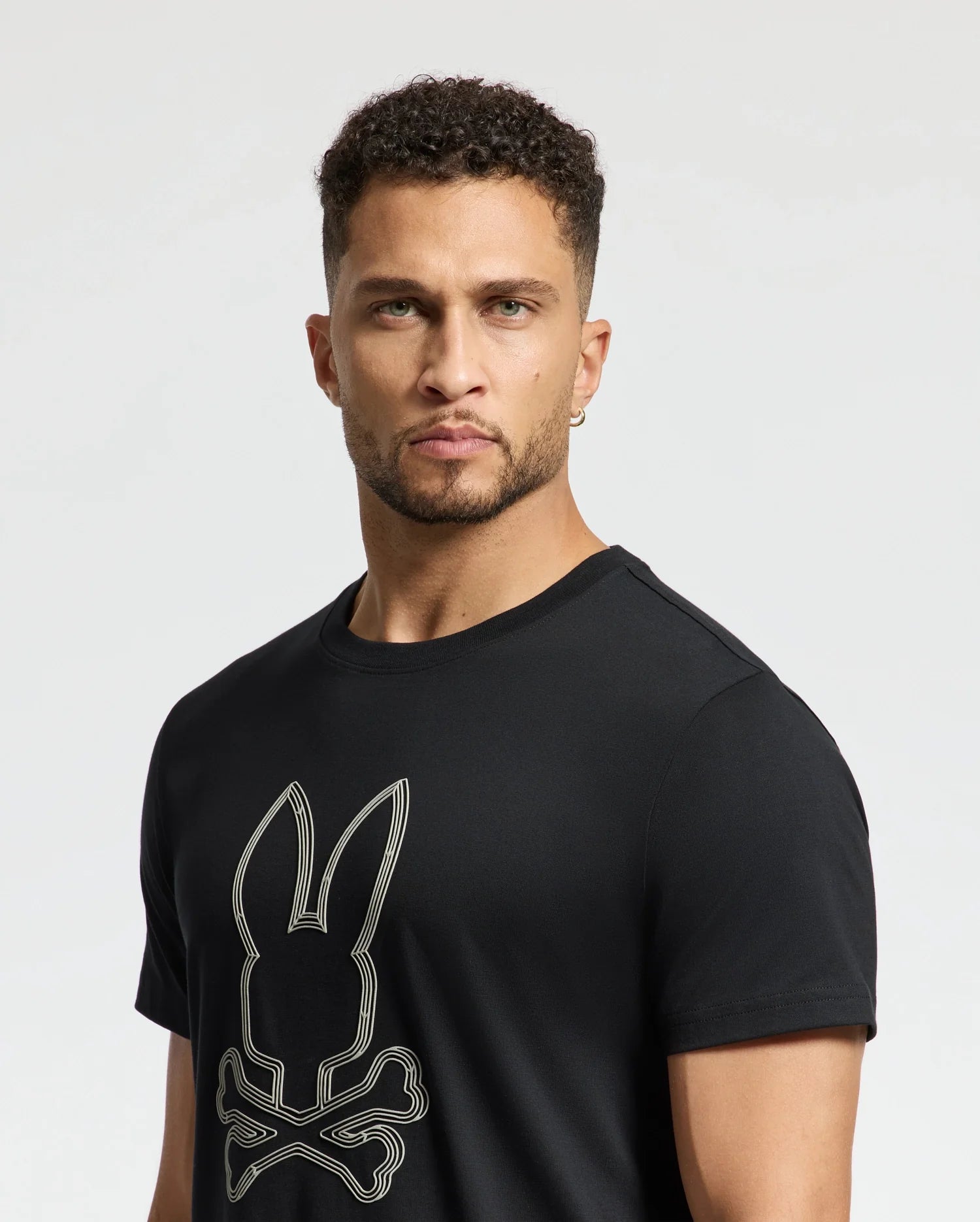 A person with short curly hair and a beard is wearing the Psycho Bunny MENS MONARC FRONT BUNNY GRAPHIC TEE (B6U211E200) in black. This T-shirt is made of 100% Pima cotton and features a rib knit crewneck, along with a stylized rabbit and crossbones design. They are looking directly at the camera against a neutral background.