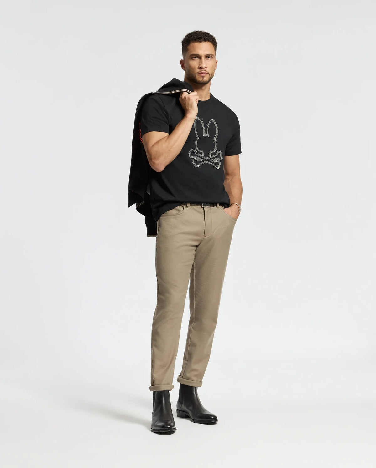 A man is wearing the Psycho Bunny MENS MONARC FRONT BUNNY GRAPHIC TEE (B6U211E200), crafted from 100% Pima cotton, along with beige pants and black boots. He has a jacket draped over his shoulder and stands confidently on a white background, showcasing the casual rib knit crewneck style of the tee.