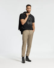 A man is wearing the Psycho Bunny MENS MONARC FRONT BUNNY GRAPHIC TEE (B6U211E200), crafted from 100% Pima cotton, along with beige pants and black boots. He has a jacket draped over his shoulder and stands confidently on a white background, showcasing the casual rib knit crewneck style of the tee.
