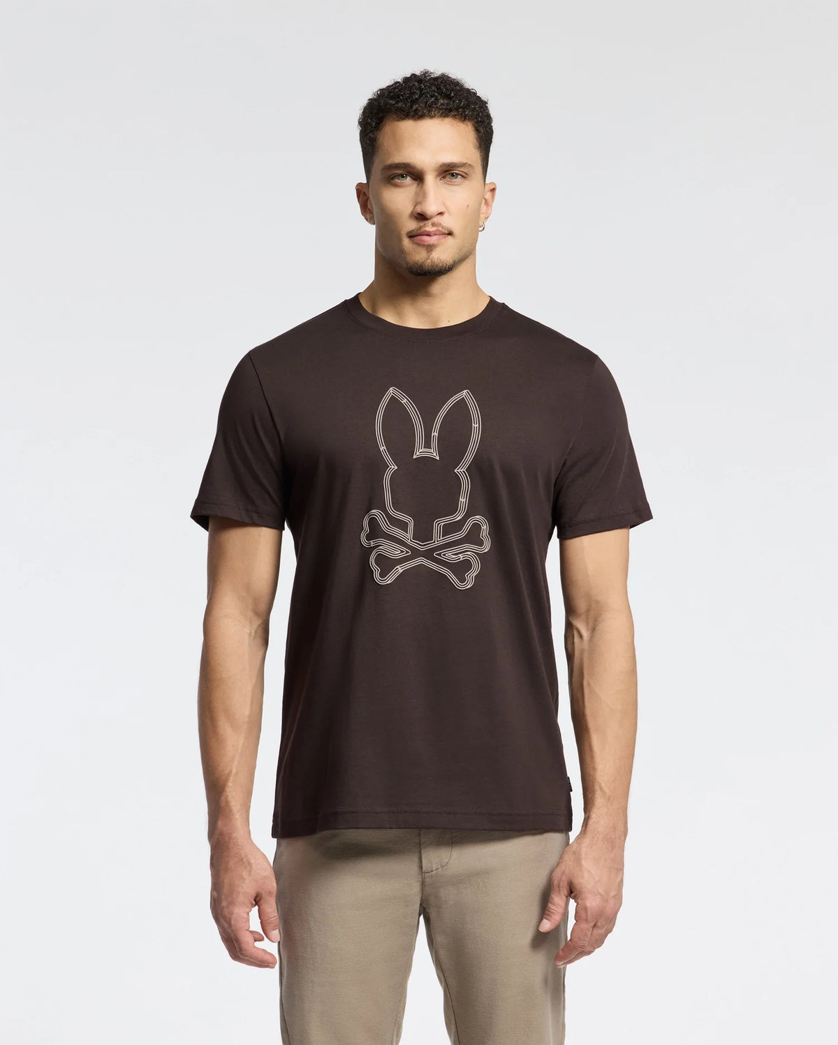 A man stands against a plain background, wearing the Psycho Bunny Men's Monarc Front Bunny Graphic Tee (B6U211E200), crafted from soft Pima cotton. The shirt showcases a raised-printed Bunny and crossbones design. His short curly hair complements his light-colored pants.