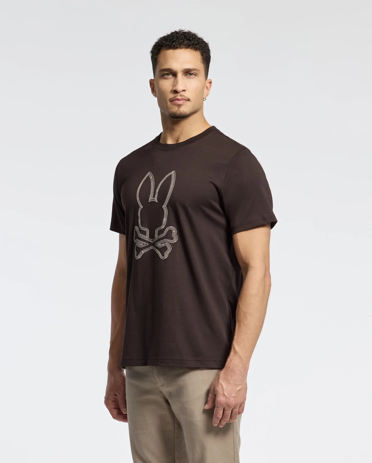 A man is wearing the Psycho Bunny MENS MONARC FRONT BUNNY GRAPHIC TEE (B6U211E200), made from Pima cotton, showcasing a raised-print Bunny with crossbones design. His short curly hair stands out against the plain white background as he gazes forward with a neutral expression.